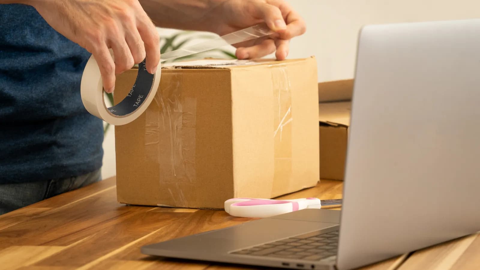 Ein Paket wird mit Klebeband verschlossen, daneben steht ein aufgeklappter Laptop